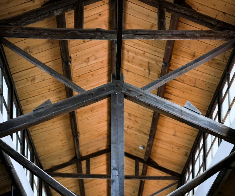 Charpente en bois vue de l'intérieur : poinçon, entraits et jambes de force sombres sous un lambris clair, entre de grandes vitres
