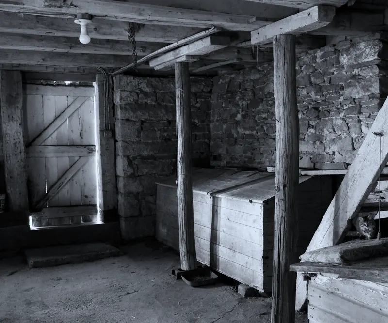 Intérieur d'une grange en pierre et en bois, en noir et blanc : solives, poteau de bois brut et porte à croix de Saint-André