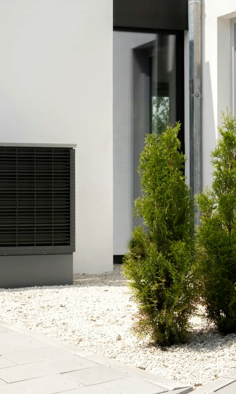 Grille d'une pompe à chaleur extérieure contre un mur blanc, entre deux thuyas et un lit de gravier clair