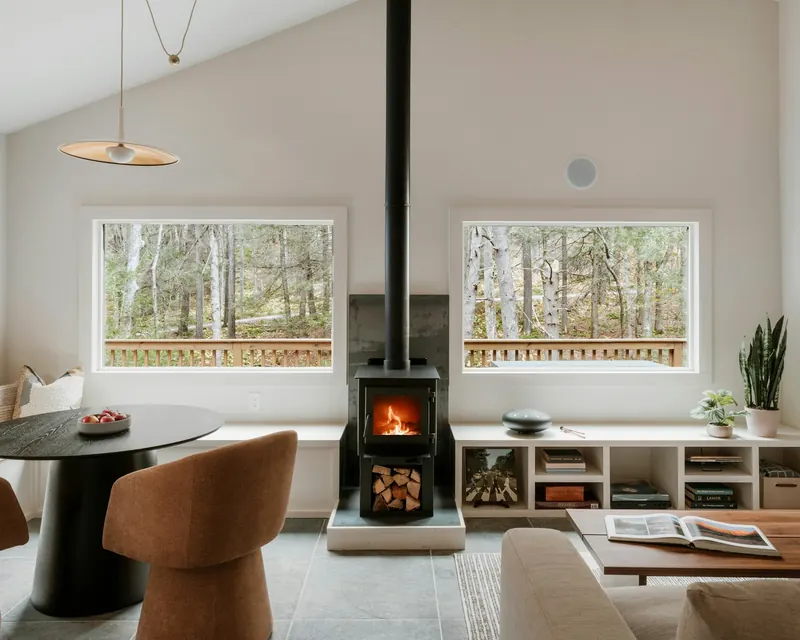 Salon clair et chaleureux : poêle à bois allumé sous une grande baie sur la forêt, fauteuil couleur terre cuite et table basse en noyer