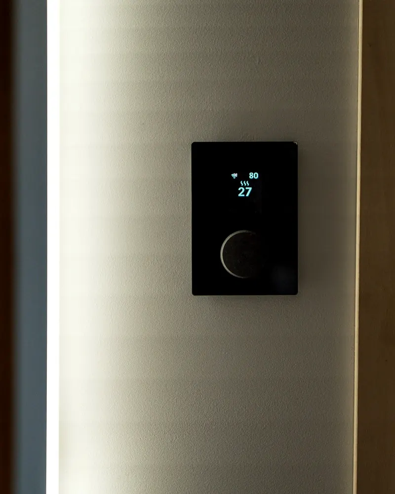 Thermostat mural noir à écran lumineux et bouton rond, posé sur un mur gris
