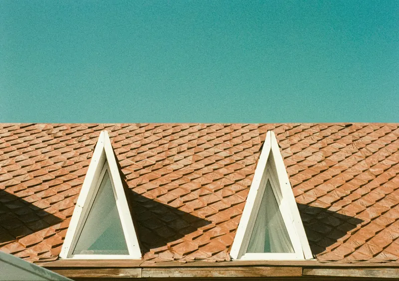 Deux lucarnes triangulaires blanches dans un pan de toit à écailles couleur terre cuite, sous un ciel sans nuage