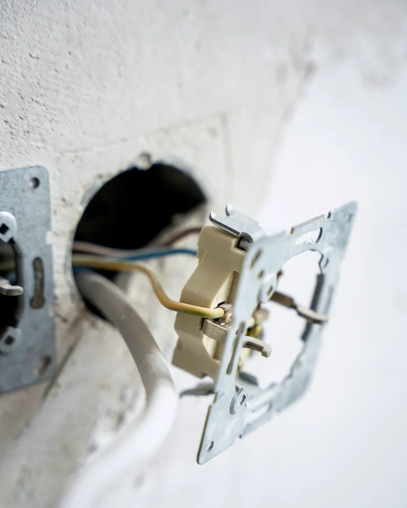 Mécanisme de prise sorti de son encastrement dans un mur blanc, avec ses fils dénudés