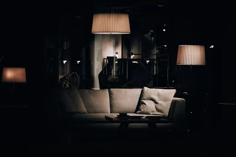 Salon plongé dans la nuit, éclairé seulement par une suspension et des lampadaires à abat-jour plissé autour d'un canapé