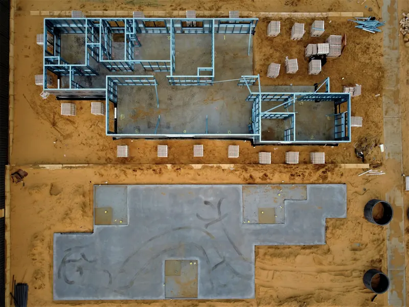 Chantier vu du ciel : ossature métallique d'une maison sur un terrain de sable orange, et dalle de béton grise à côté