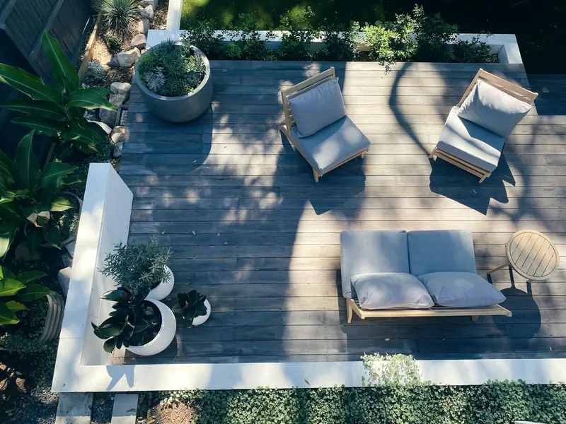 Vue de dessus d'une terrasse en bois grisé avec fauteuils et canapé de jardin, pots de plantes et muret blanc, entourée de massifs