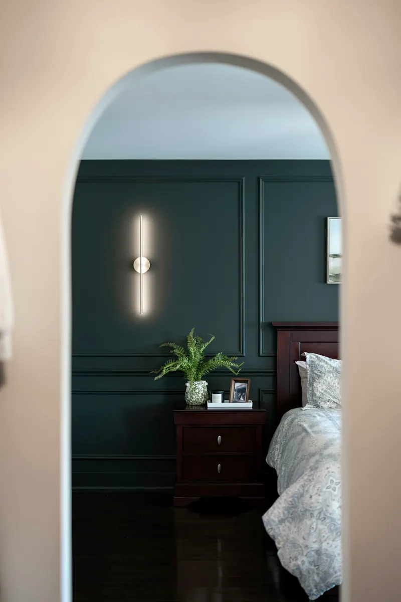 Chambre vue par une arche : mur vert profond à moulures, applique lumineuse, table de chevet et lit