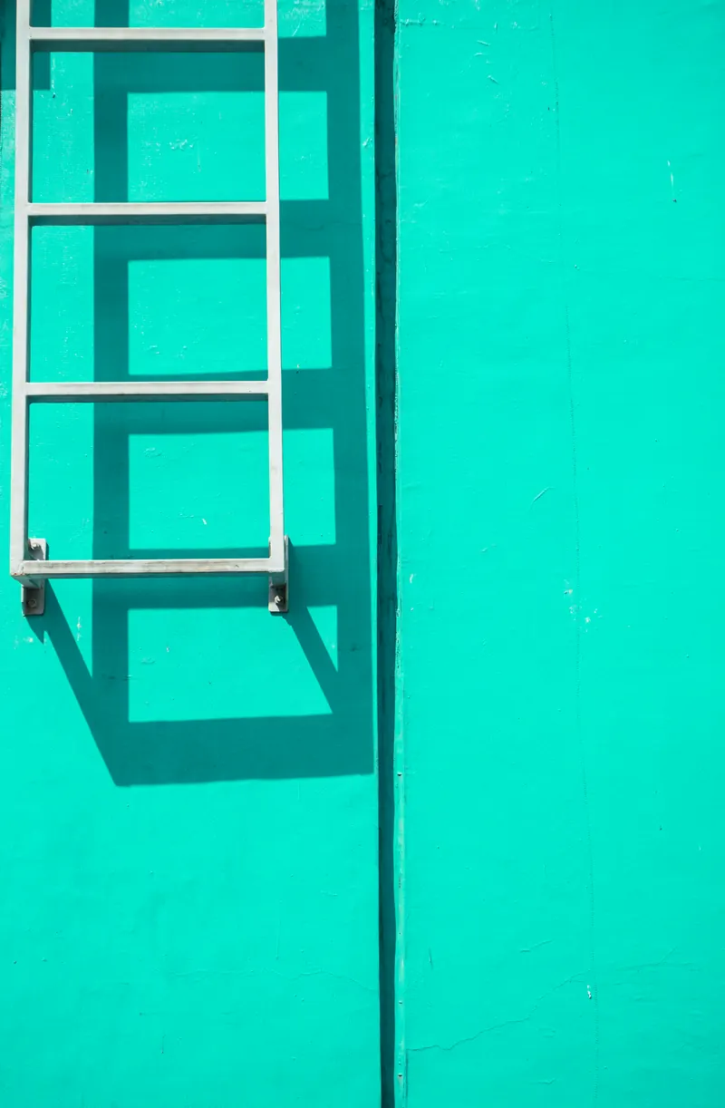 Échelle blanche fixée contre un grand mur peint en vert menthe, son ombre dessinée sur l'enduit