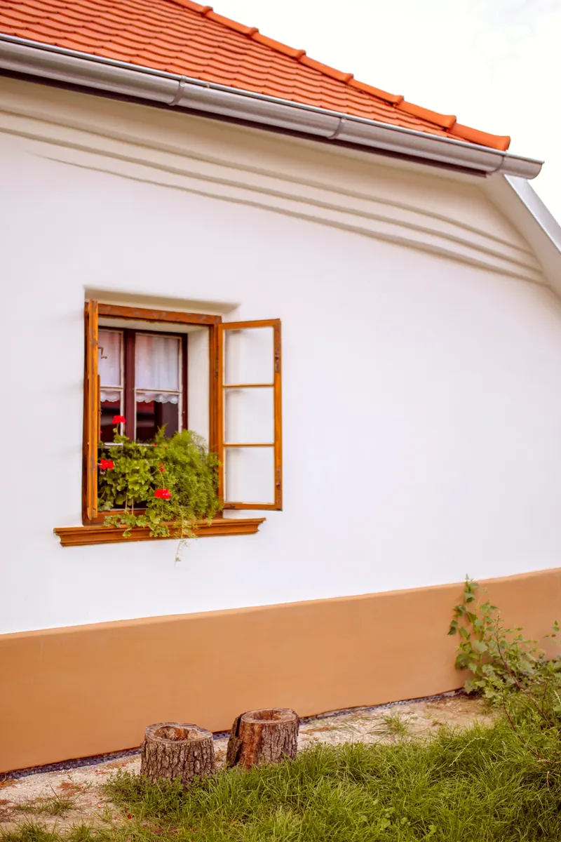 Façade enduite en blanc avec un soubassement ocre, une fenêtre en bois fleurie et un toit de tuiles