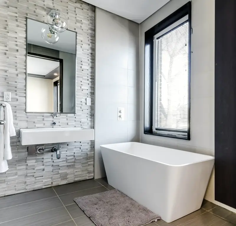 Baignoire blanche aux lignes droites sous une haute fenêtre, vasque suspendue devant un mur de pierre en lamelles grises