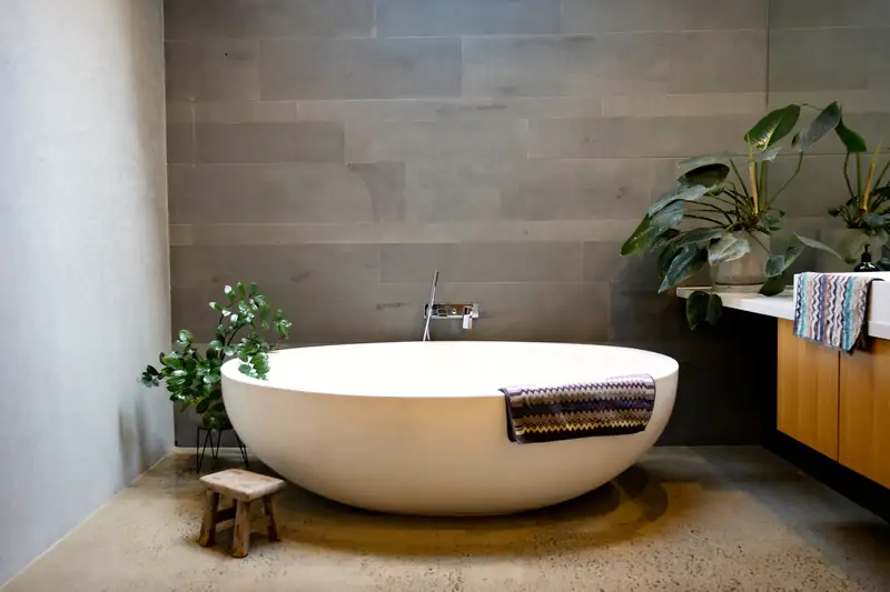 Grande baignoire ovale blanche devant un mur gris, sol en béton poli et plantes vertes