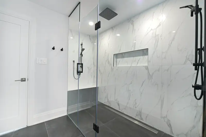Douche vitrée aux grands carreaux d'aspect marbre blanc veiné de gris, robinetterie noire et caniveau au sol